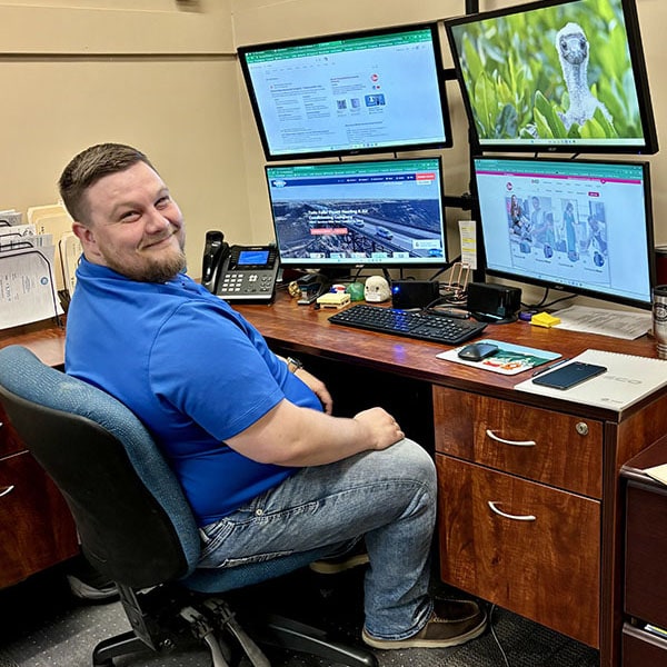 A man in a blue polo shirt and jeans sits in a blue office chair at a wooden desk. He is smiling and looking over his shoulder at the camera. His workstation includes four computer monitors displaying various content, a keyboard, mouse, phone, and other office supplies.