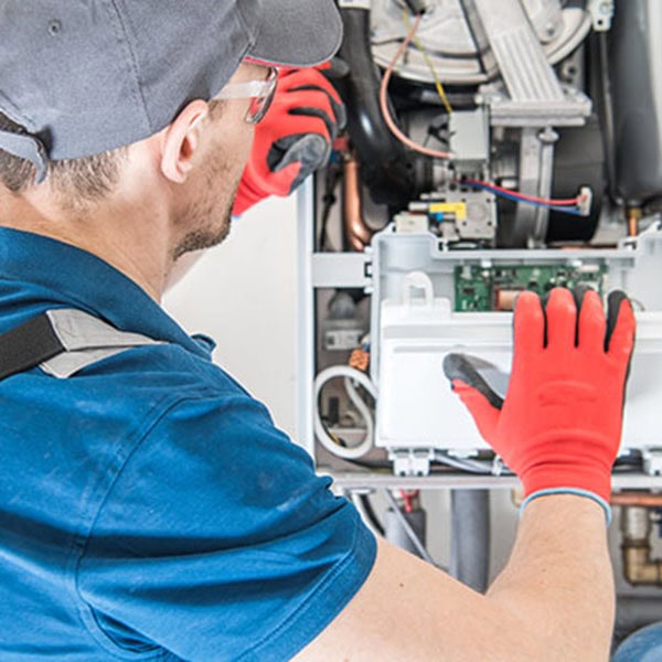 A technician wearing a blue shirt and red gloves works on installing a gas furnace, surrounded by tools and equipment.
