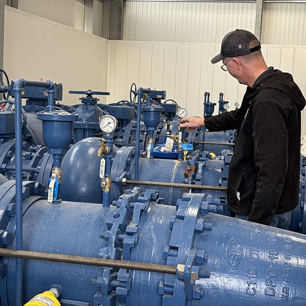 A technician wearing a black jacket and baseball cap is examining a complex network of large blue pipes, valves, and pressure gauges in an industrial setting. He is pointing at one of the gauges.