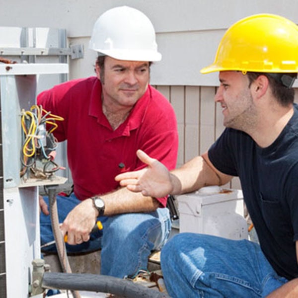 Two men wearing hard hats converse about air conditioner repair beside a heat pump
