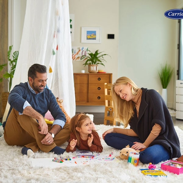 A family plays on a fluffy rug in a living room with a Carrier air purifier visible in the background, suggesting a focus on indoor air quality.