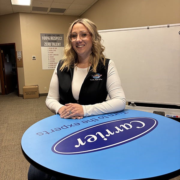 Smiling woman seated at a round table with a Carrier logo, inside a professional office space. She wears a branded vest for Terry’s Heating & Air Conditioning, representing radon testing and indoor air quality services.