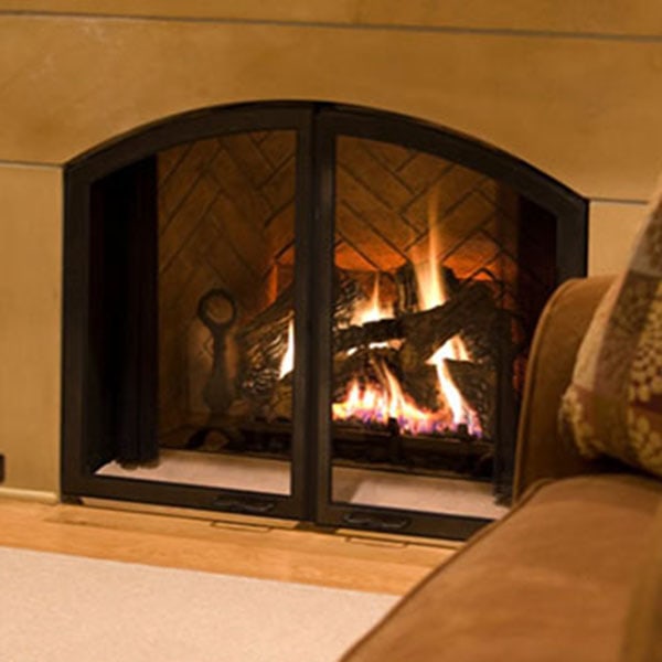 A lit fireplace with glass doors next to a brown couch with patterned pillows in a cozy living room setting.