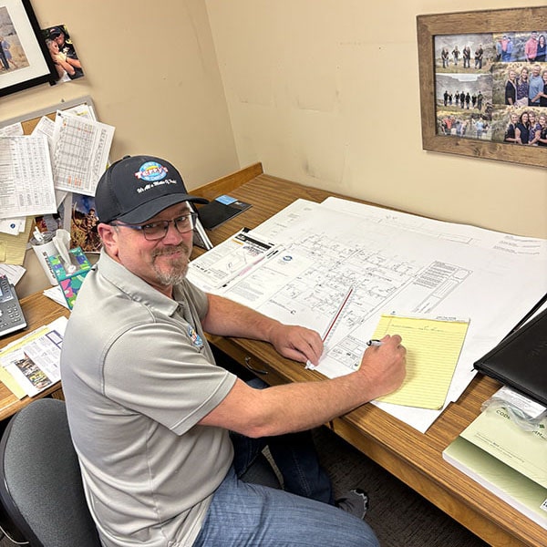 Zone Control Systems 1_600x600 A smiling man wearing a baseball cap and glasses sits at a wooden desk covered with papers, blueprints, and office supplies. He is holding a pen and yellow notepad, appearing to be reviewing a large technical drawing labeled "Zone Control System." The background shows a beige wall with framed photos and other papers.